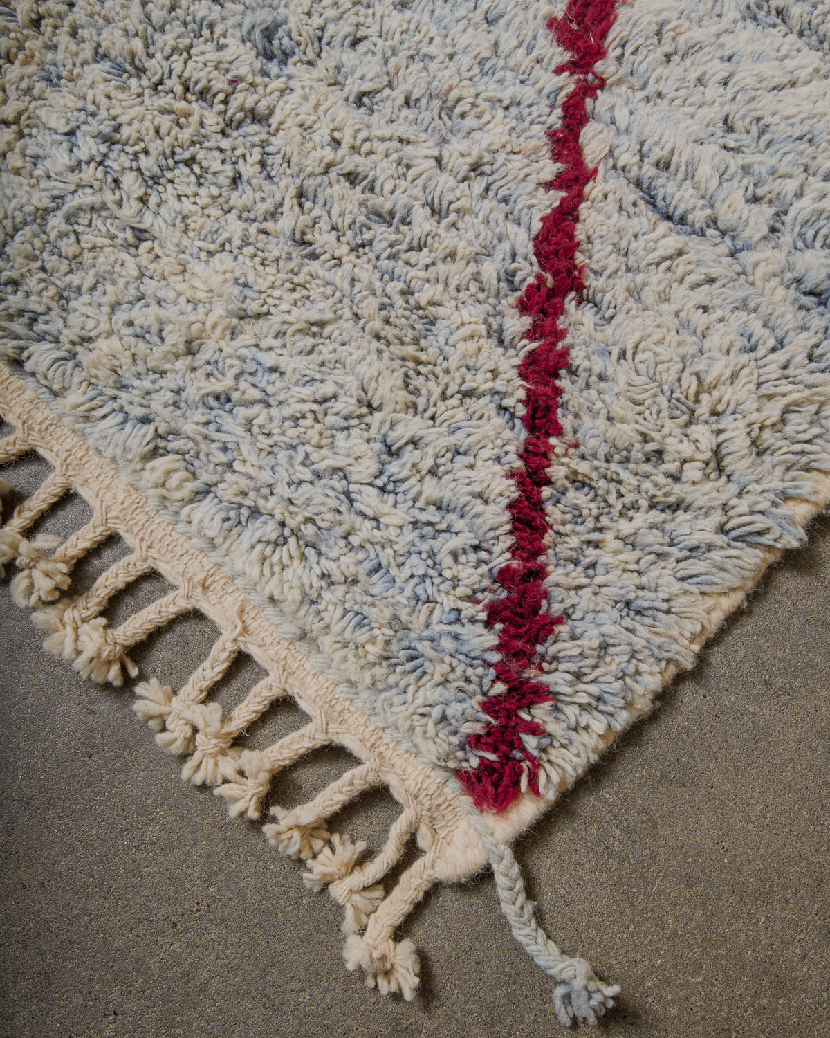 Close-up of a textured rug with a red stripe and tassels on a neutral background