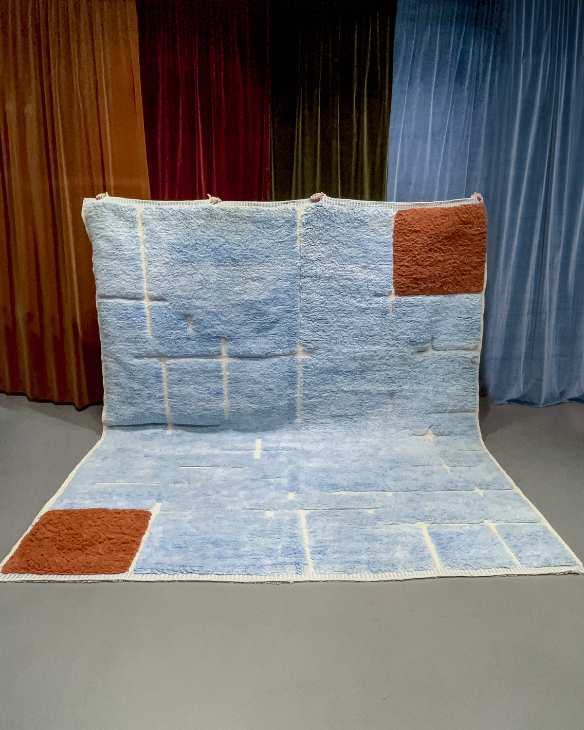 Large sky blue Berberlin M'rirt with brown squares at two corners being held up in front of velvet curtains.