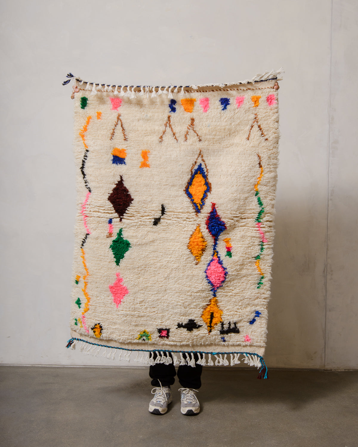 Berberlin Azilal rug in cream wool with colourful cotton  being held up in front of a white wall.