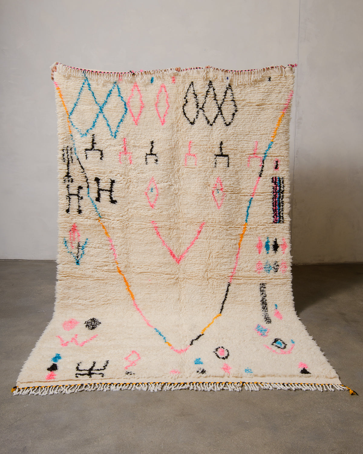 Berberlin Azilal rug made of fluffy cream wool and colourful design, held up in front of a white wall and cement floor.