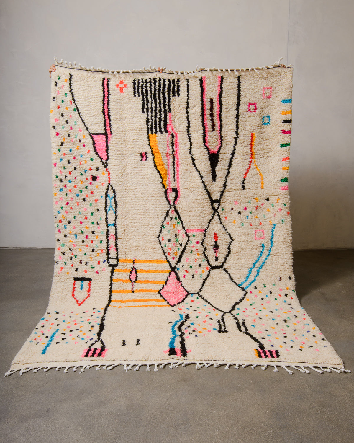 Berberlin Azilal rug made of fluffy cream wool and colourful design, held up in front of a white wall and cement floor.