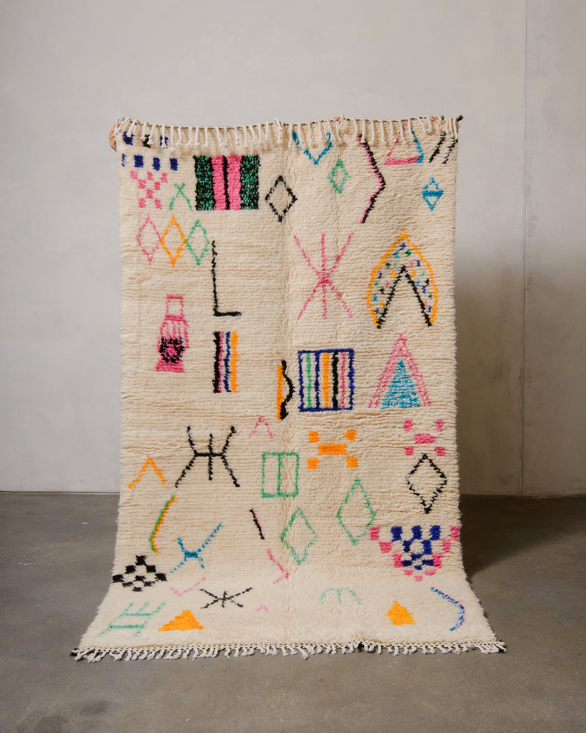 Berberlin Azilal rug made of fluffy cream wool and colourful design, held up in front of a white wall and cement floor.