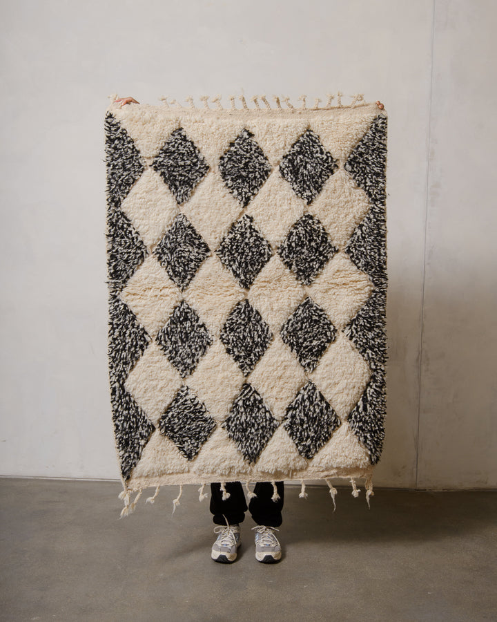 Small Berberlin Beniourain rug made of wool in black and white, being held up in front of a white wall. 