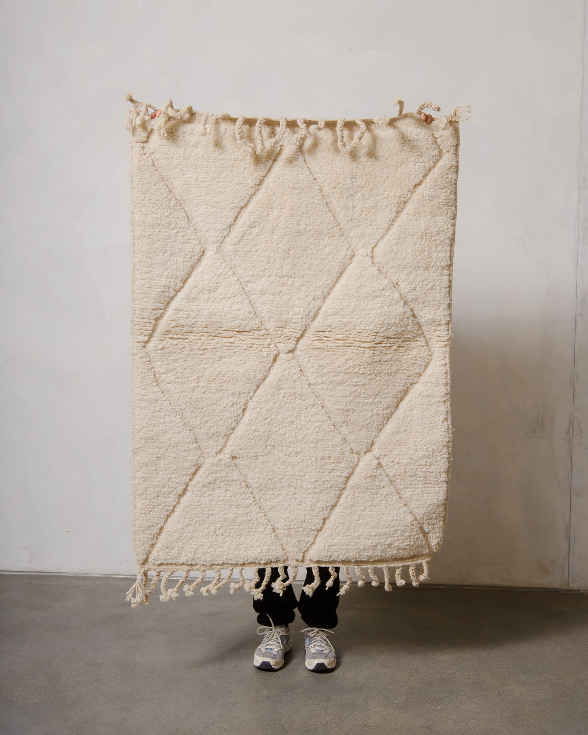 Small Berberlin Beniourain rug in cream wool with diamond design, being held up in front of a white wall. 