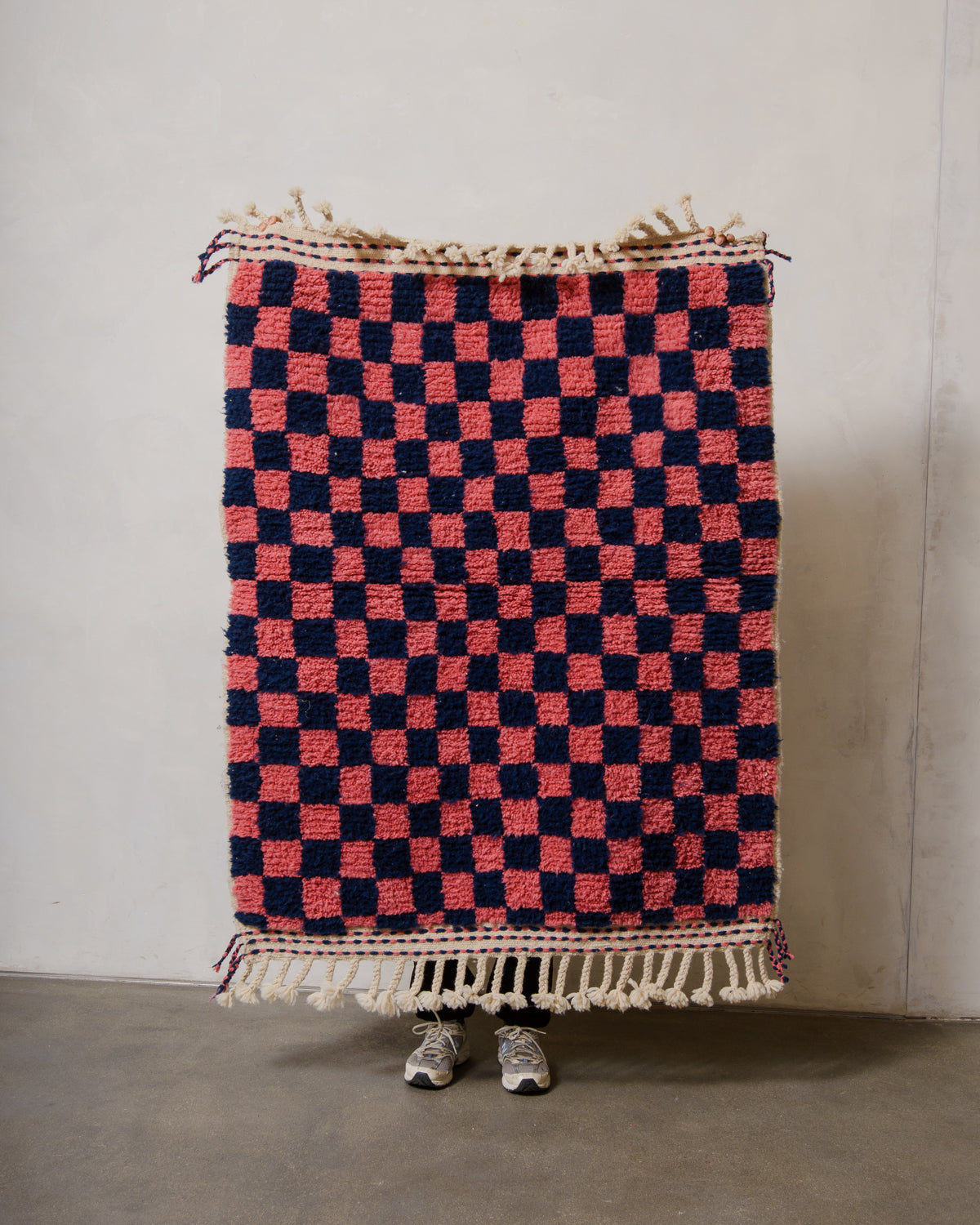 Small Berberlin Beniourain rug made of wool inl pink and dark blue checkers, being held up in front of a white wall. 