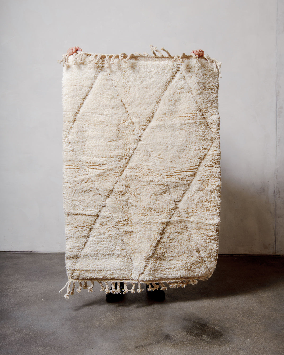 Small Berberlin Beniourain rug in cream wool with diamond design, being held up in front of a white wall. 