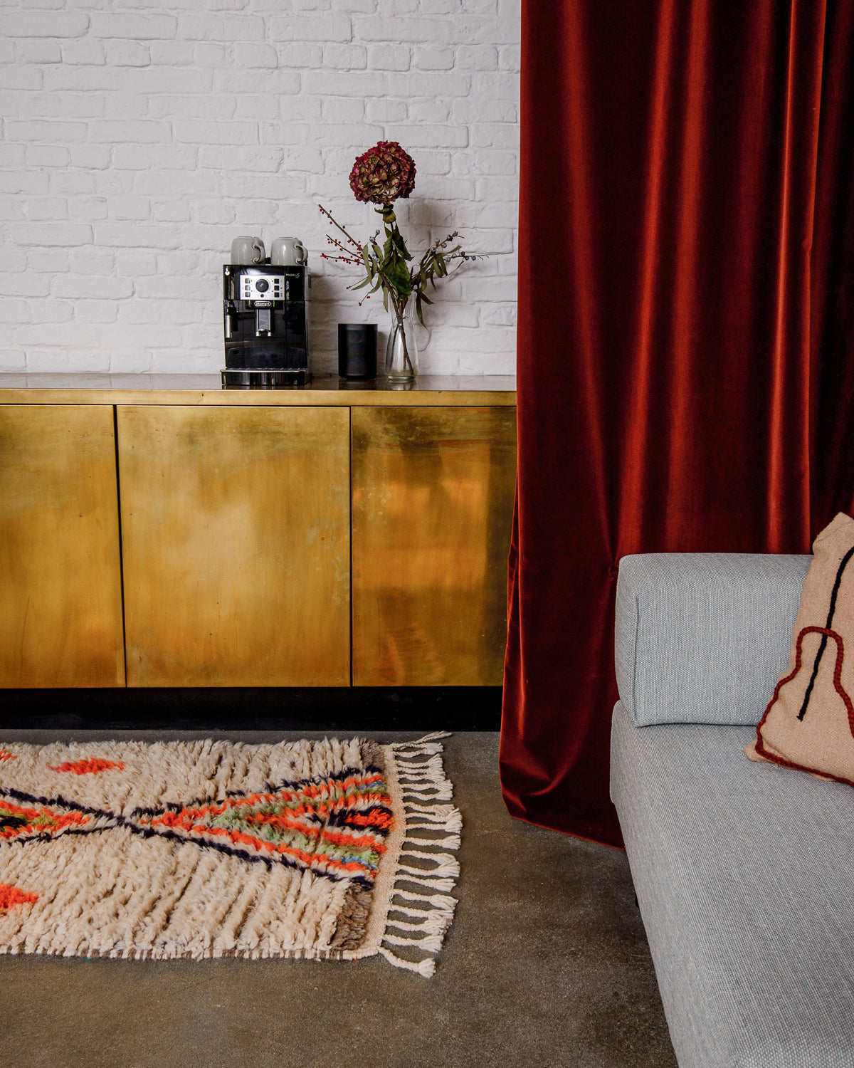 Interior with vintage runner rug on floor in front of a gold cabinet. To the right side is a red velvet curtain and the edge of a grey modular sofa.