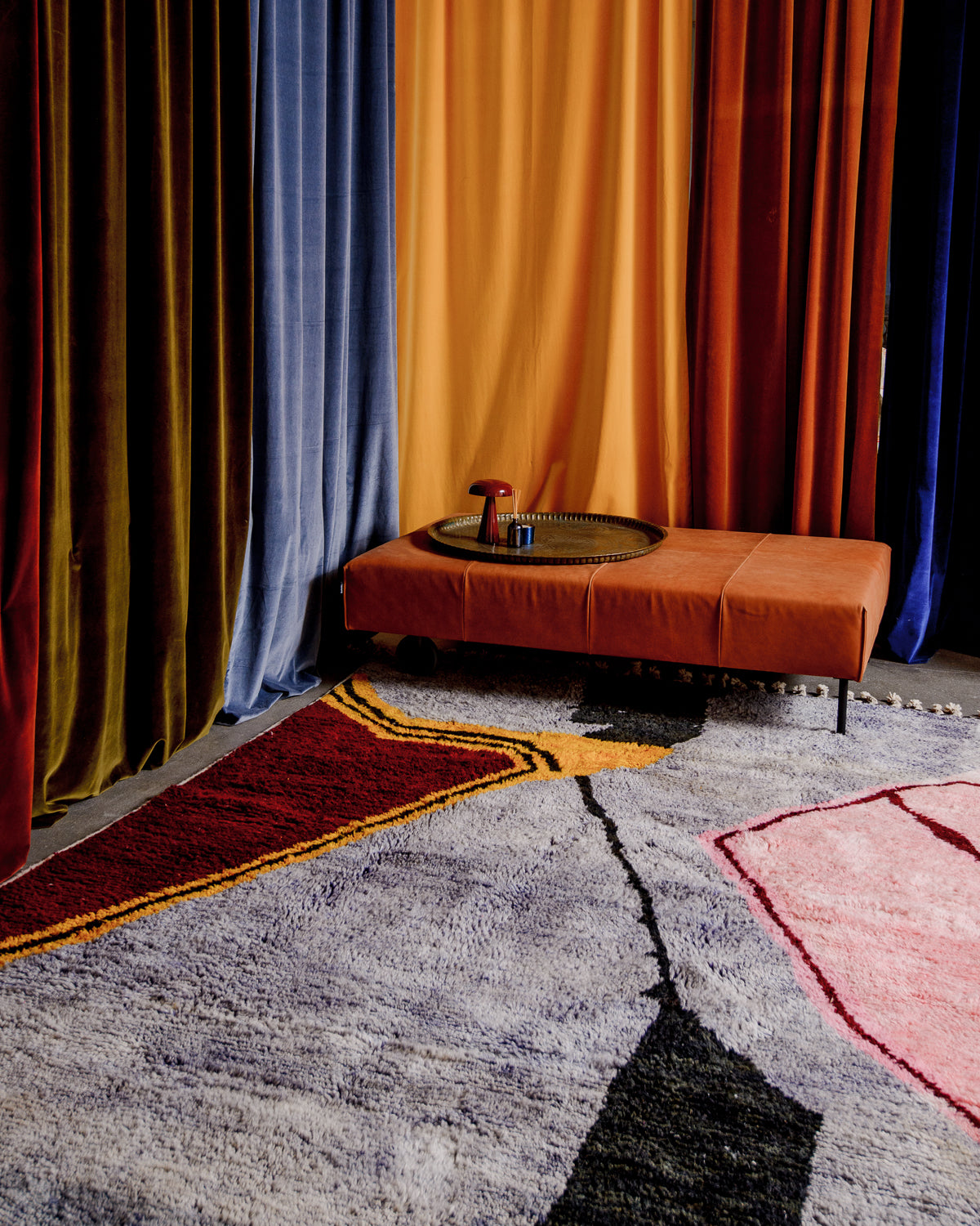 Large, colourful rug in lavender, burgundy, pink and yellow surrounded by velvet curtains in different colours and a brown leather ottoman.