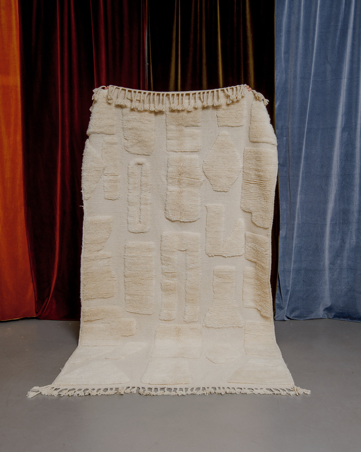 Cream wool rug with different forms in thick wool pile and a flat woven base being held up in front of velvet curtains. 