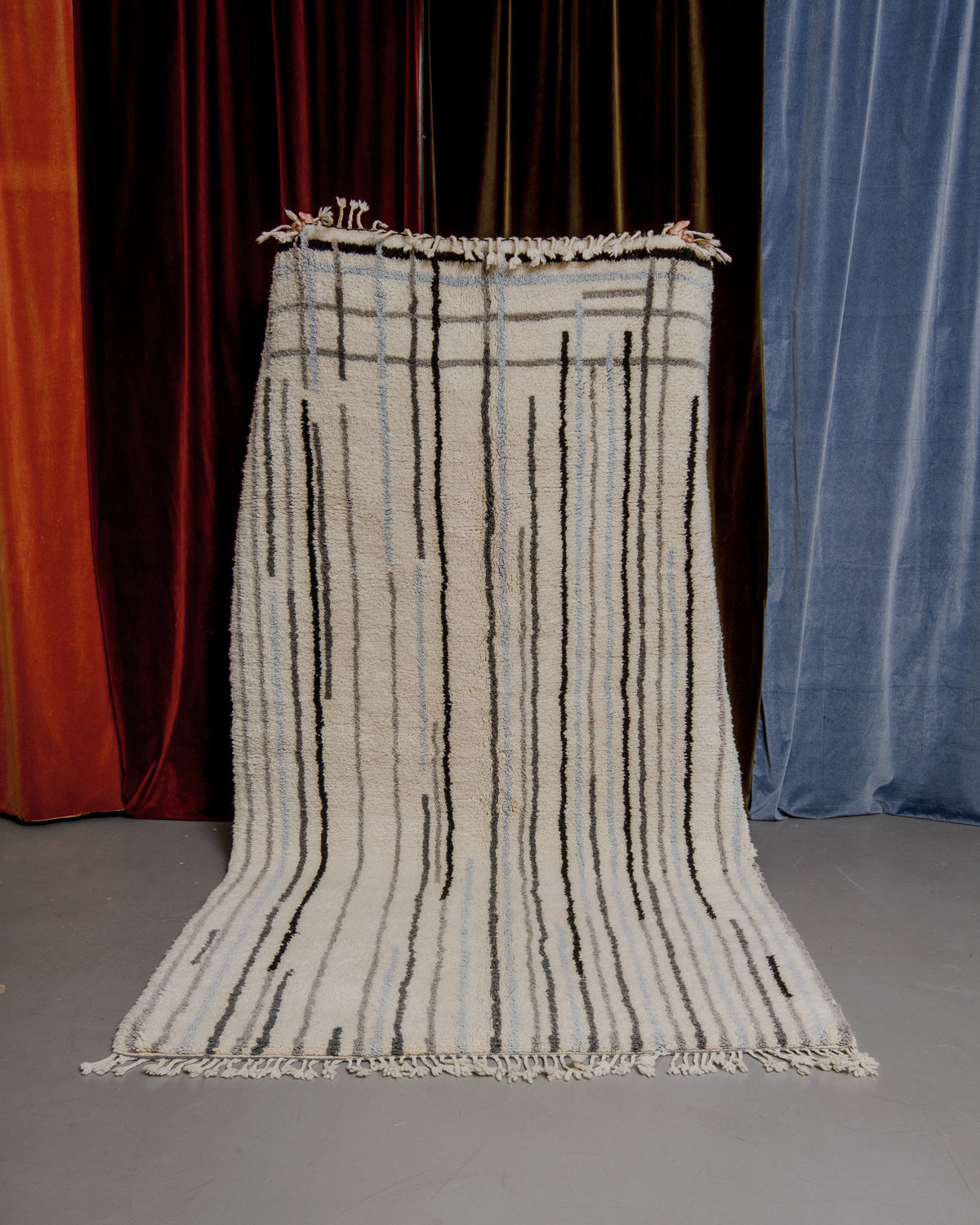 Berberlin Finest Rug made of heavy wool pile with grey, blue and black lines, being held in front of velvet curtains.