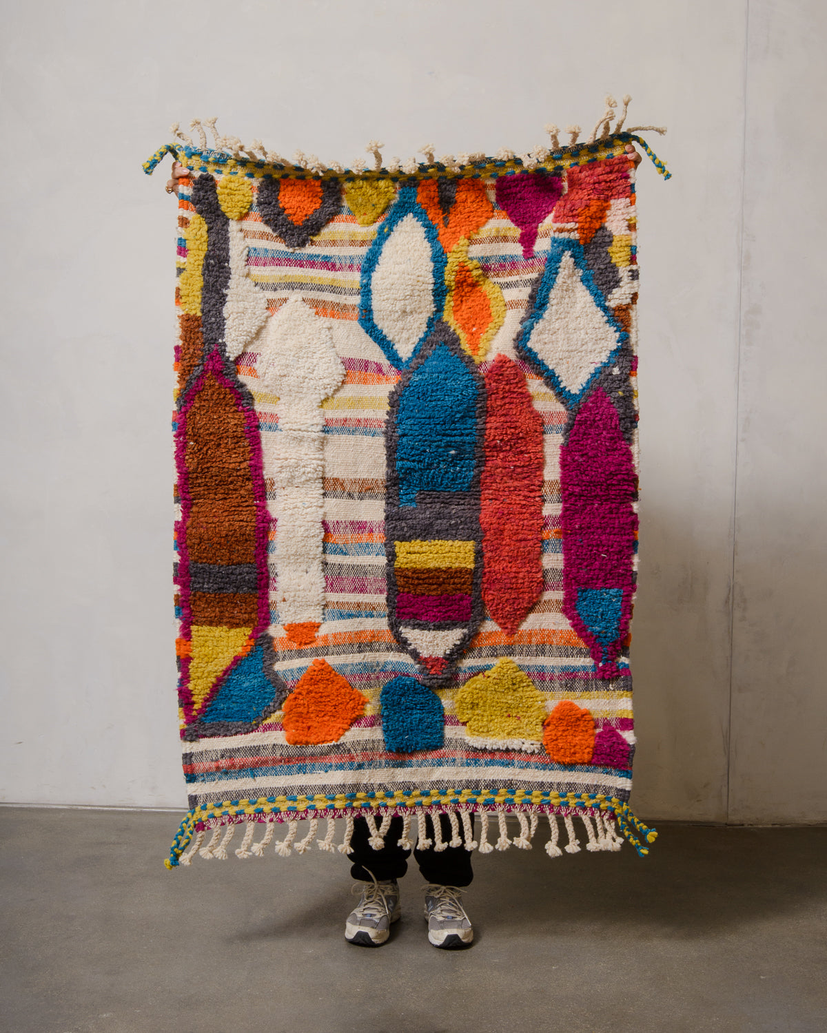 Small Berberlin Beniourain rug made of colourful wool with knotted and woven areas, being held up in front of a white wall. 