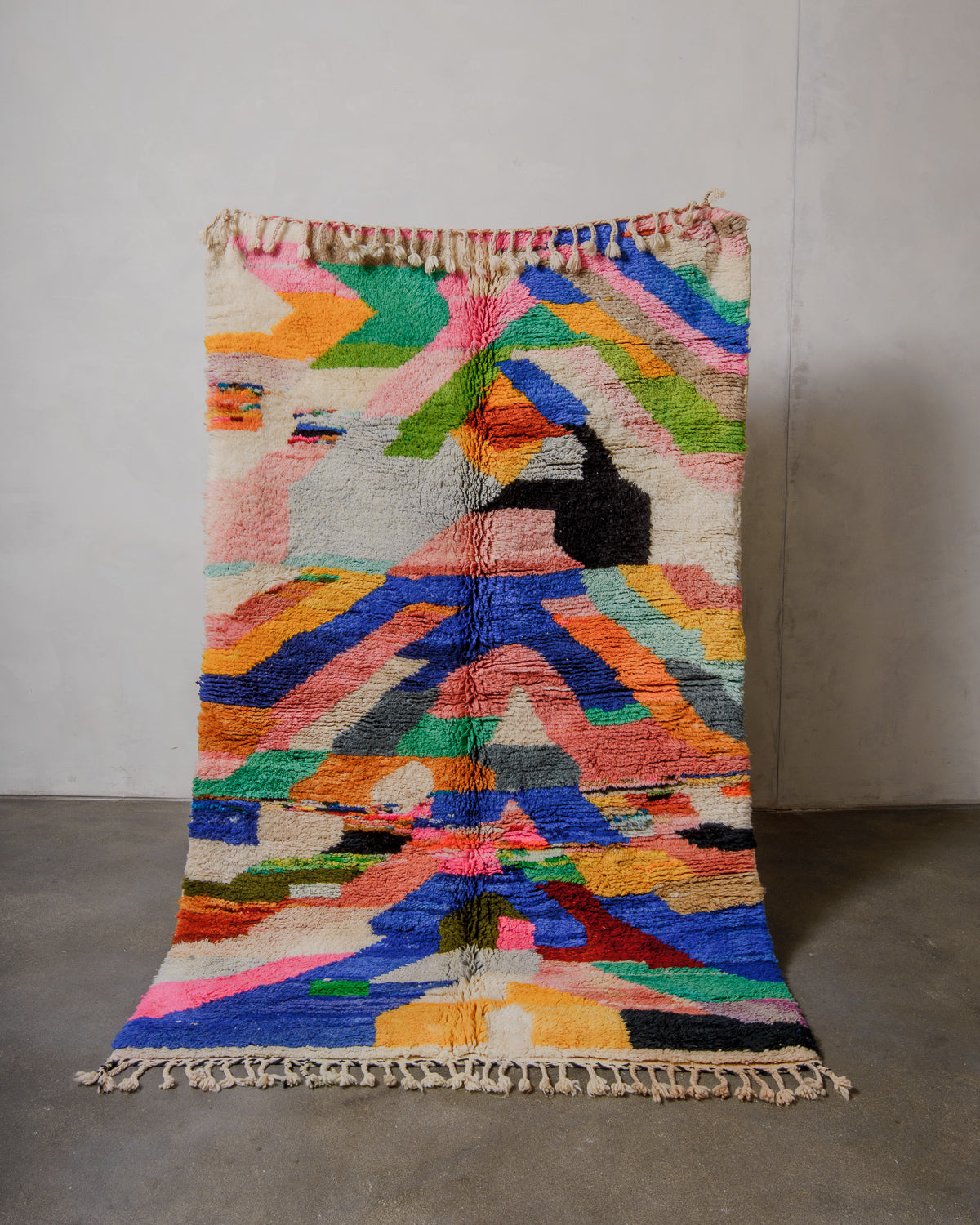 Boldly coloured wool rug with tassels being held up in front of a plain wall. 