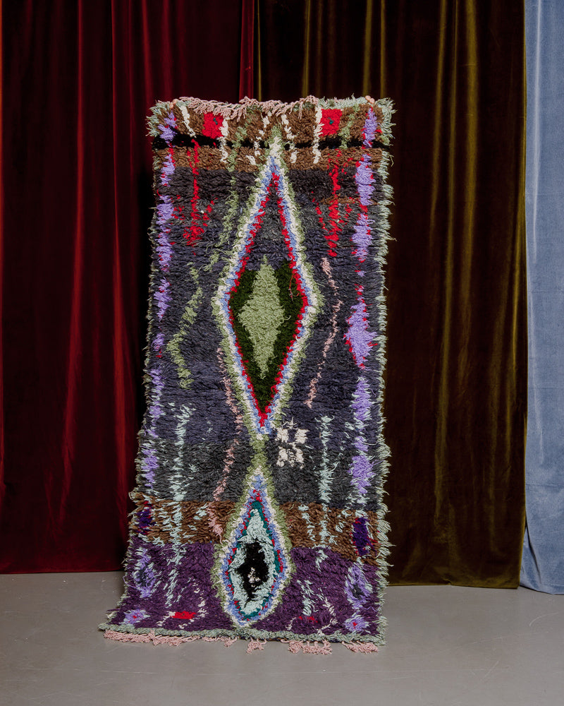 Vintage Berberlin boucherouite runner rug being held up in front of a velvet curtain. 