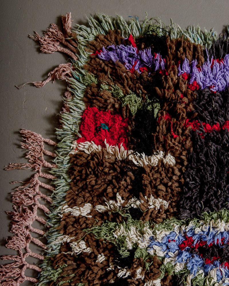 Corner of boucherouite rug with red, sage green, blue, charcoal and white cotton and, brown wool, with rose coloured tassels.