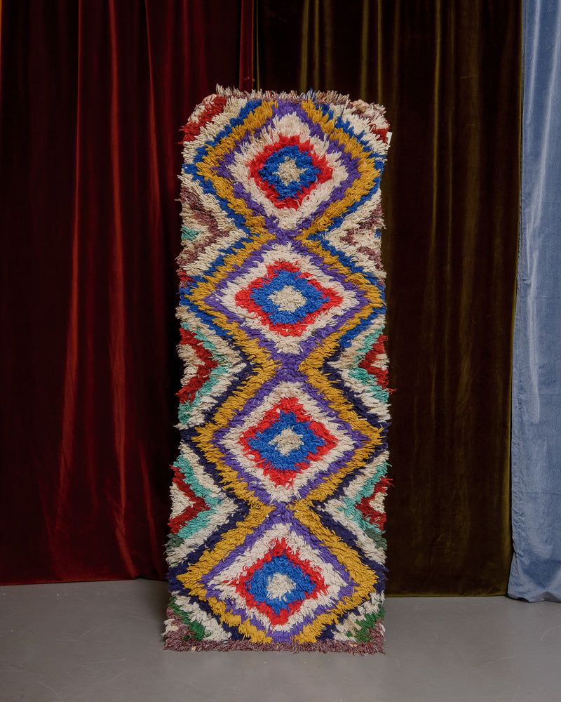 Vintage Berberlin boucherouite runner rug being held up in front of a velvet curtain. 