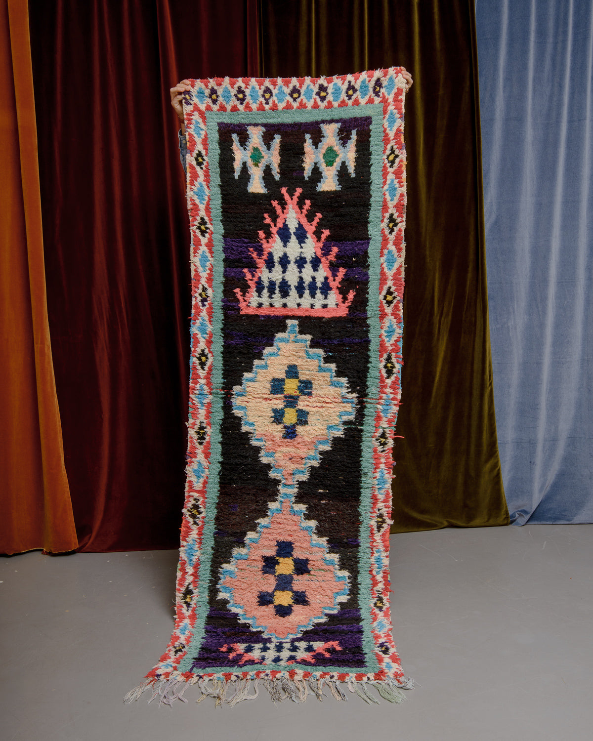 Vintage Berberlin boucherouite runner rug being held up in front of a velvet curtain. 