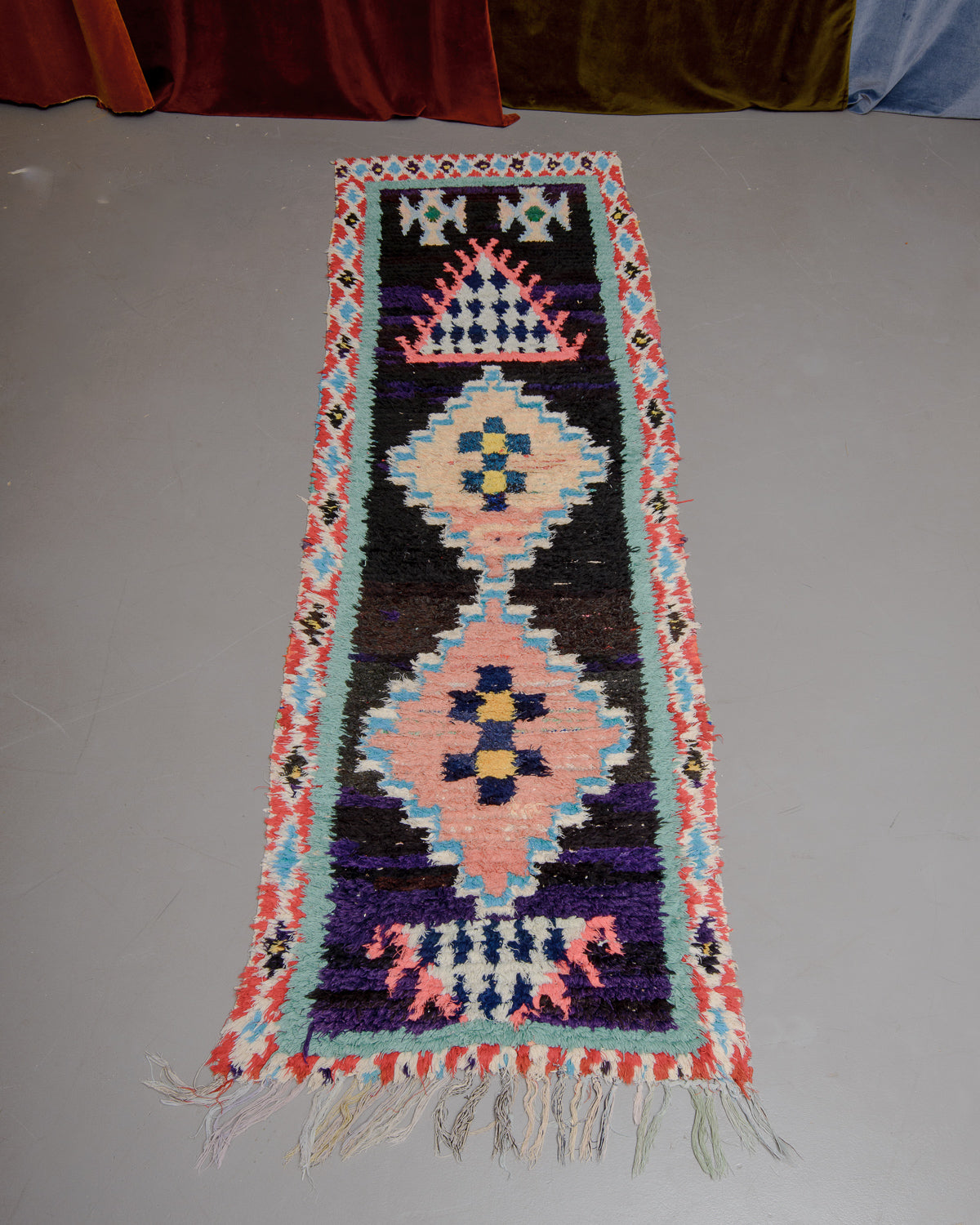Vintage Berberlin boucherouite runner rug on grey floor in front of velvet curtain. 