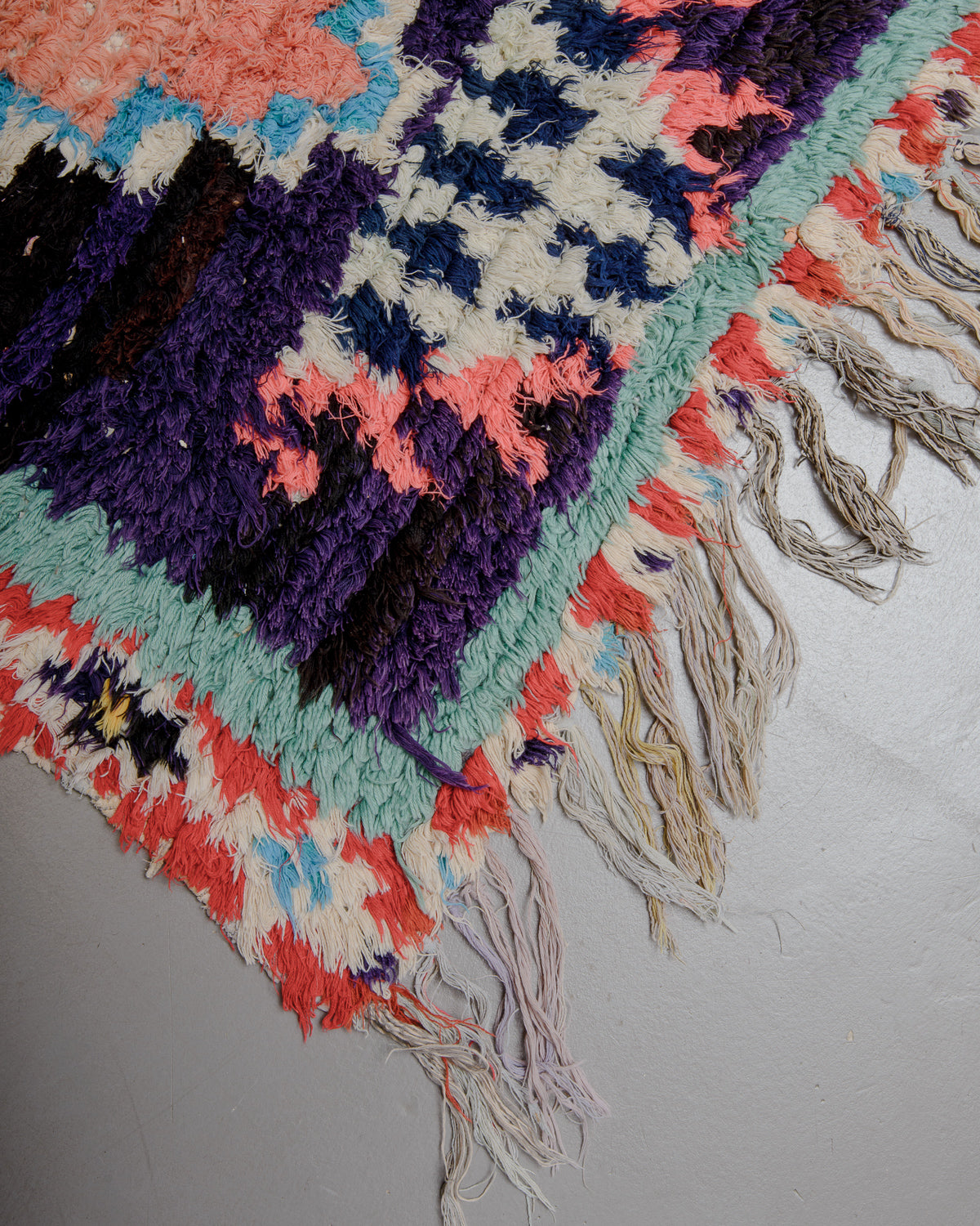 Corner of rug with purple, turquoise, red, white and blue with a fringed edge on a grey floor.