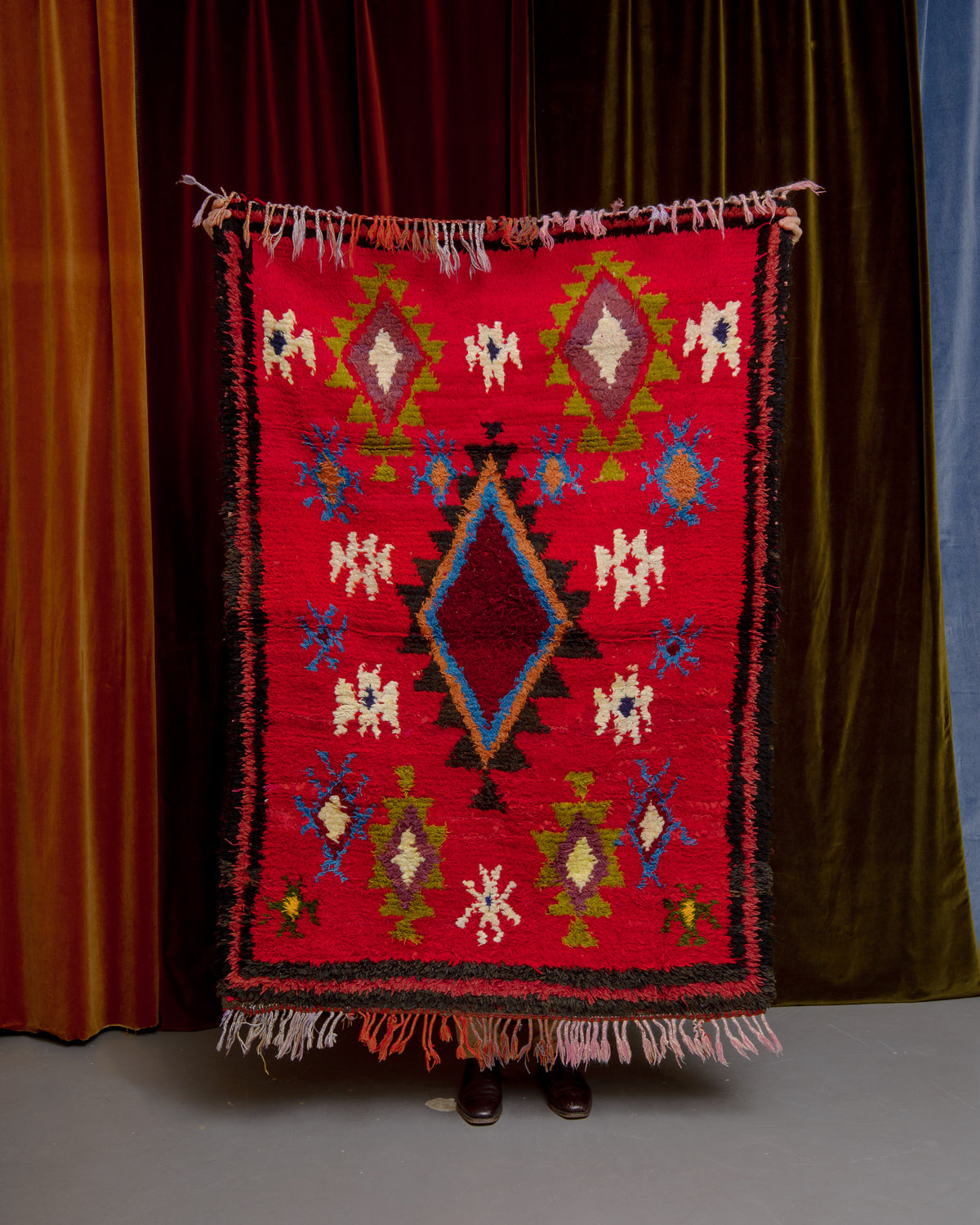 Vintage Berberlin boucherouite runner rug being held up in front of a velvet curtain. 