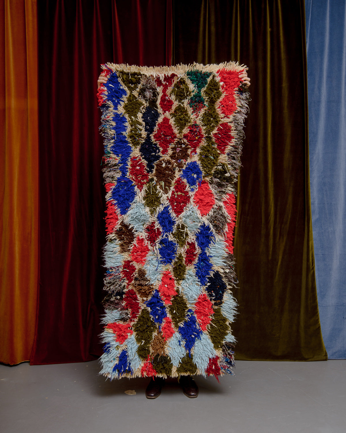 Vintage Berberlin boucherouite runner rug being held up in front of a velvet curtain. 