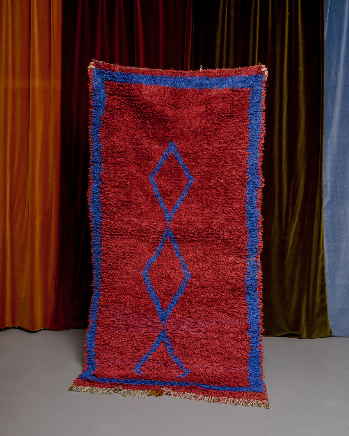 Vintage Berberlin boucherouite runner rug being held up in front of a velvet curtain. 