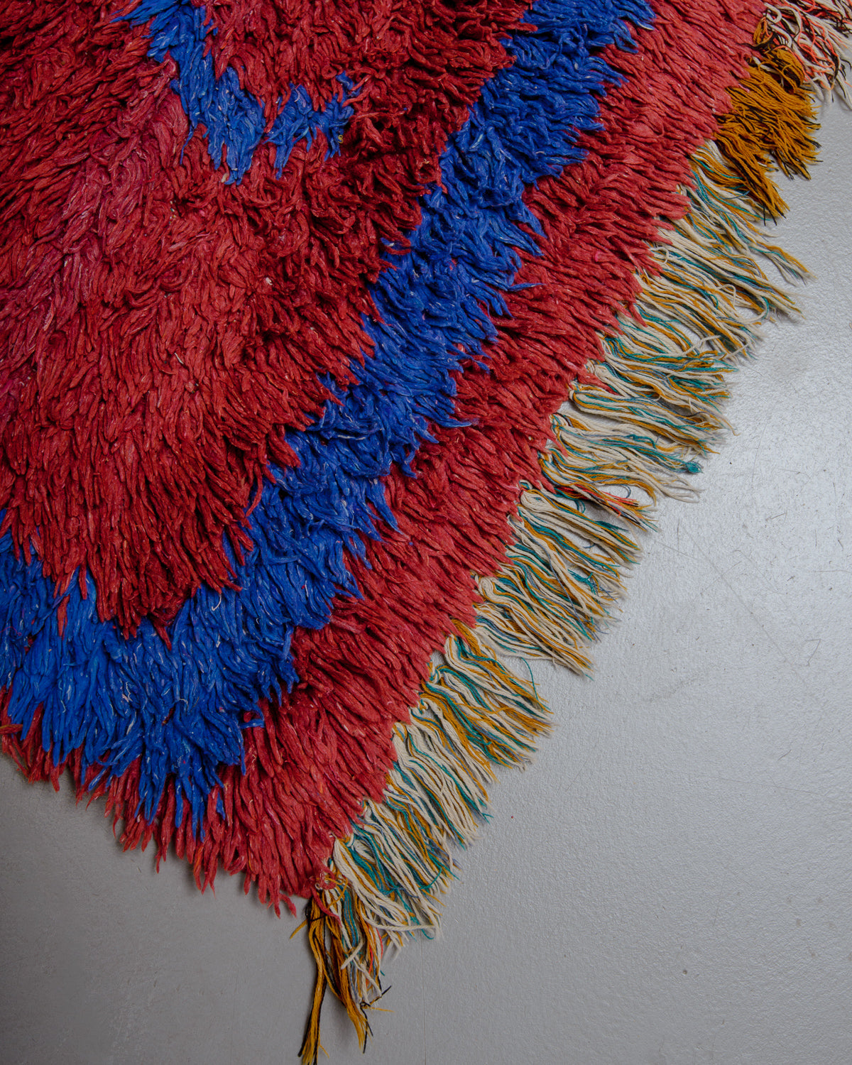 Corner of rust red and blue rug with multicoloured cotton tassels.