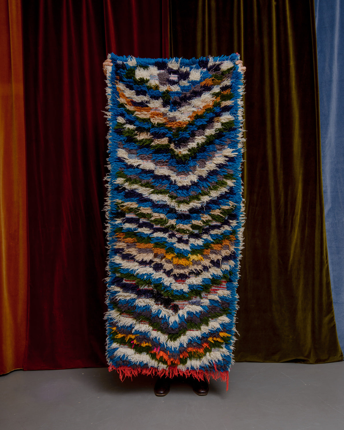 Vintage Berberlin boucherouite runner rug being held up in front of a velvet curtain. 