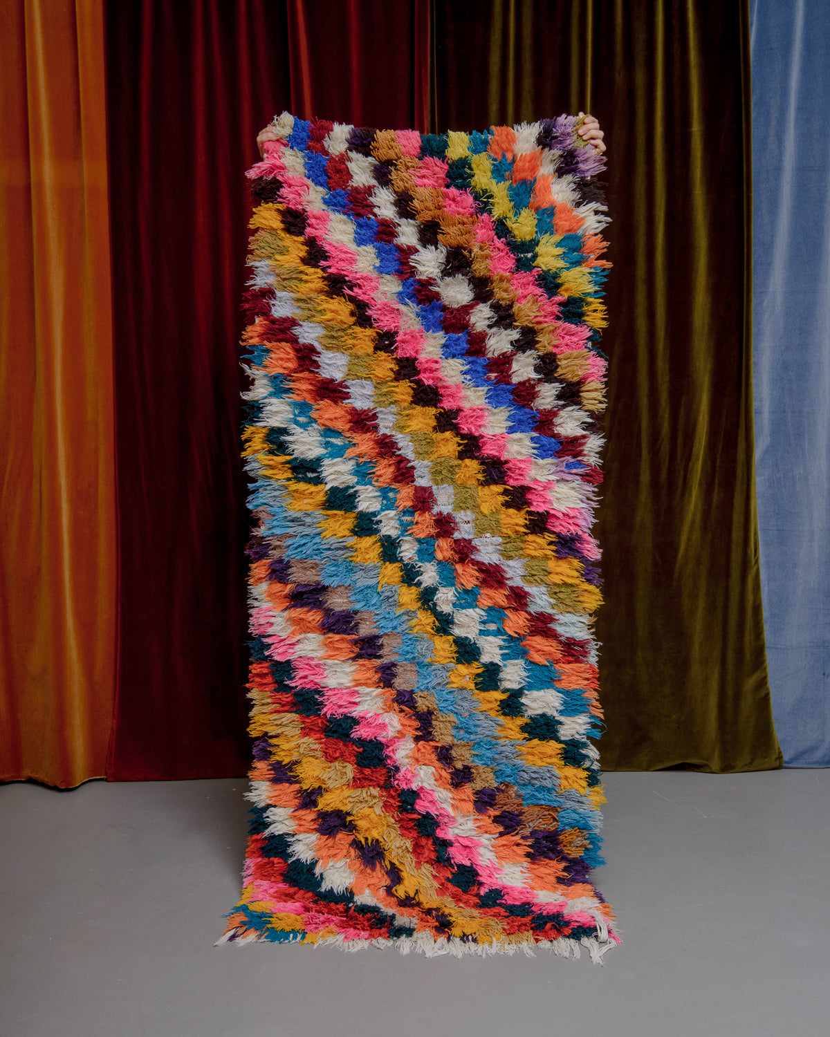 Vintage Berberlin boucherouite runner rug being held up in front of a velvet curtain. 