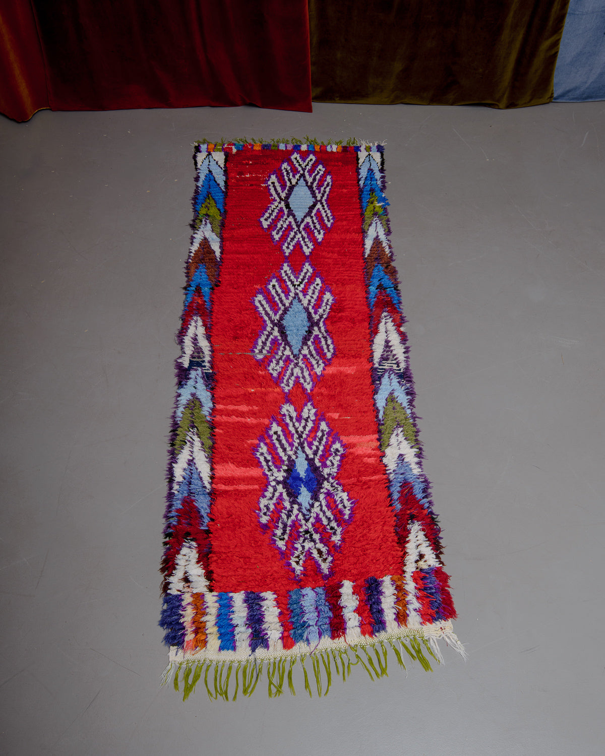 Vintage Berberlin boucherouite runner rug on grey floor in front of velvet curtain. 