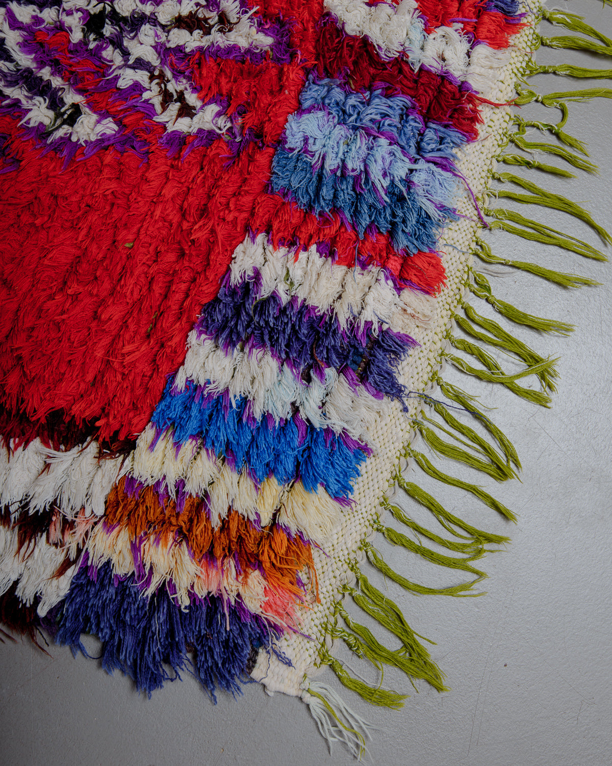 Corner of boucherouite rug with red, purple, blue and white cotton with green cotton tassels.