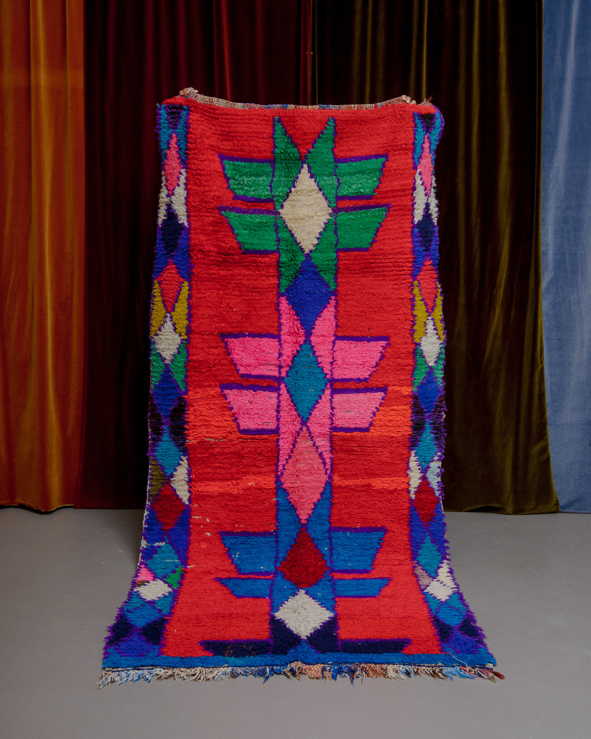 Vintage Berberlin boucherouite runner rug being held up in front of a velvet curtain. 
