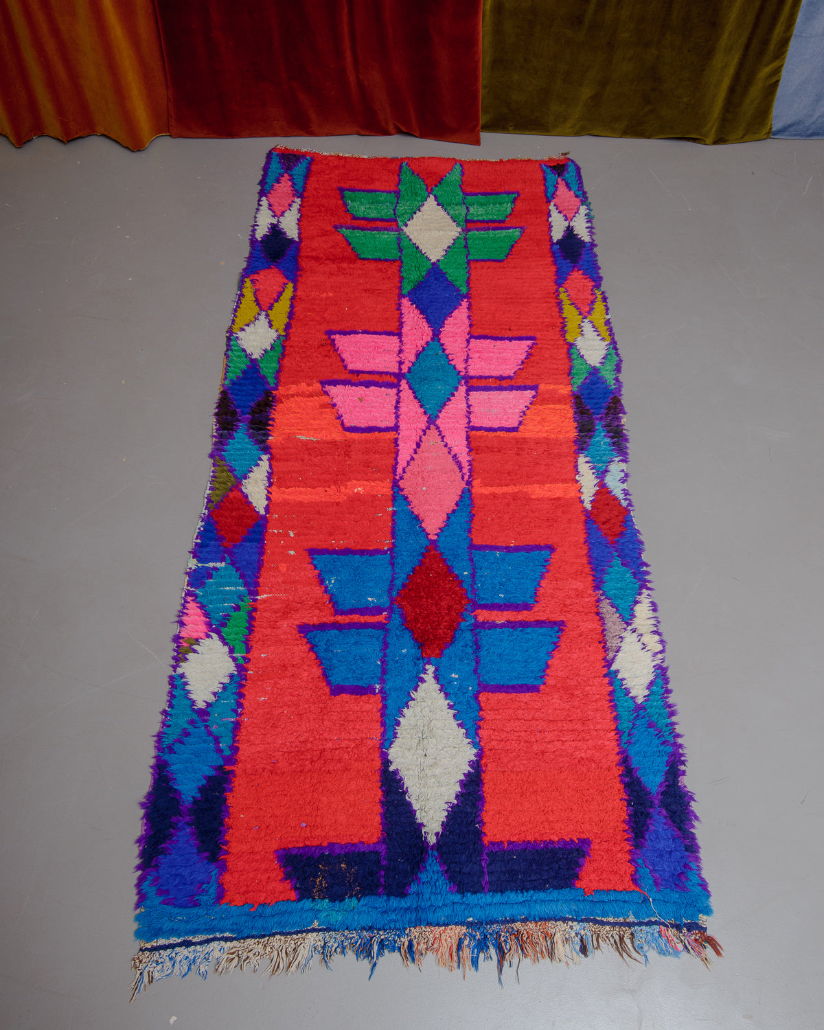 Vintage Berberlin boucherouite runner rug  on grey floor in front of velvet curtain. 