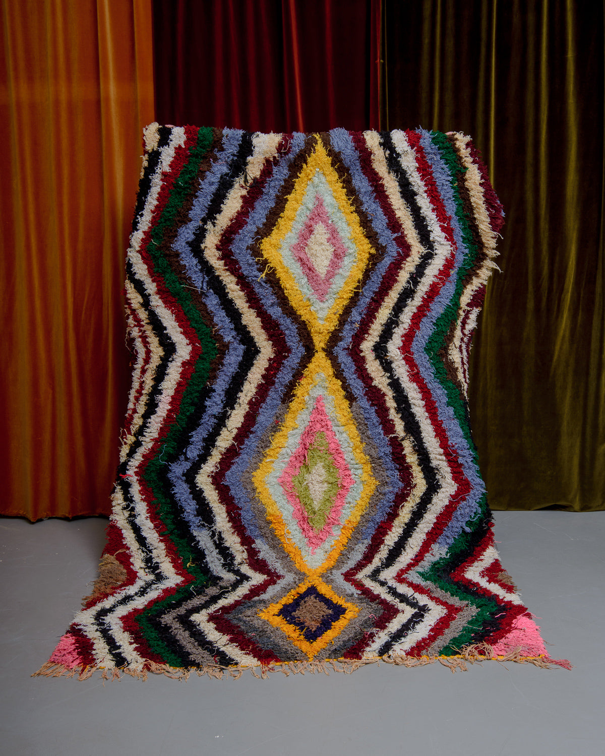 Boucherouite rug being held up in front of velvet curtains.