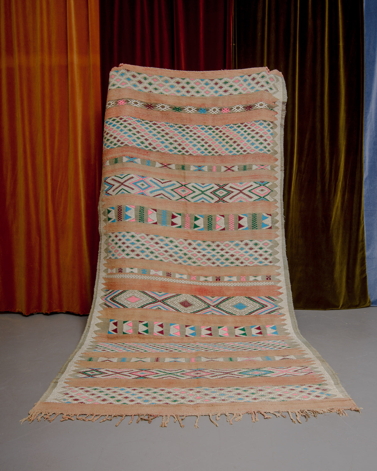 Vintage Kelim rug in muted shades of beige, rose, green, blue and pink being held in front of velvet curtains. 