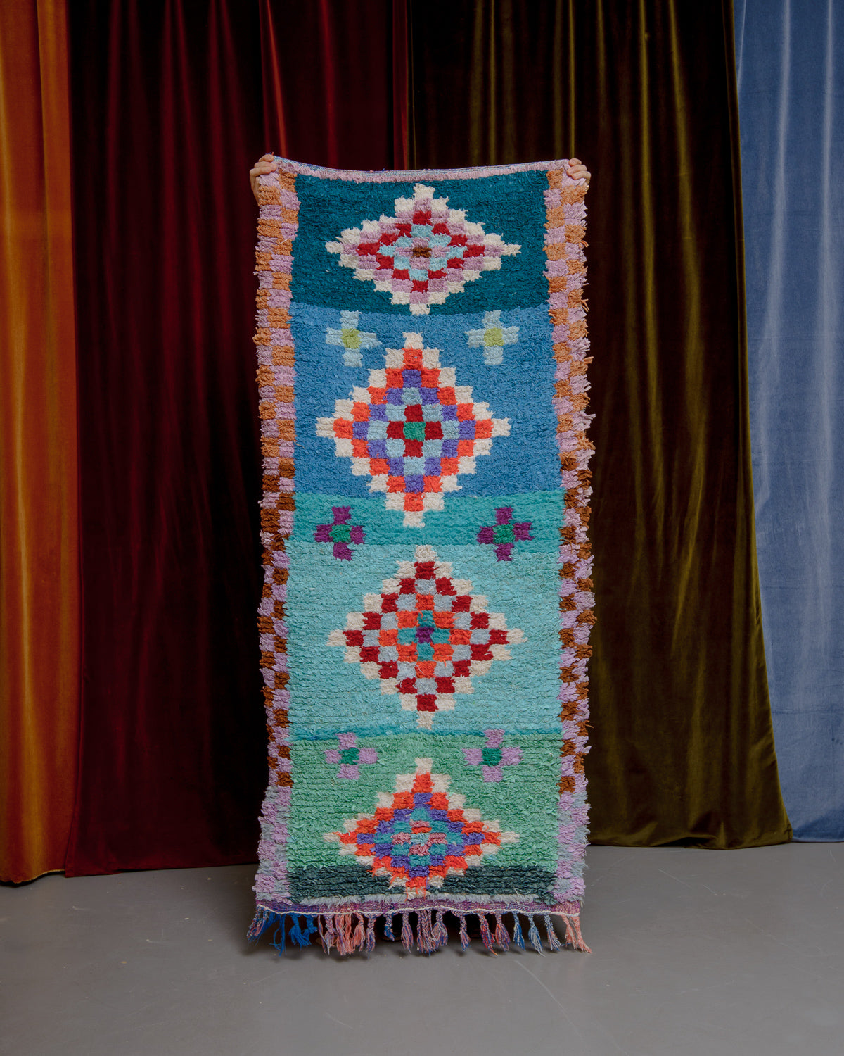 Vintage Berberlin boucherouite runner rug being held up in front of a velvet curtain. 