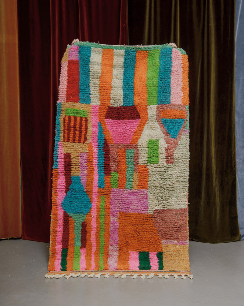 Vintage Boujaad rug made of blue, green, pink, orange and yellow stripes and shapes being held in front of a velvet curtain.