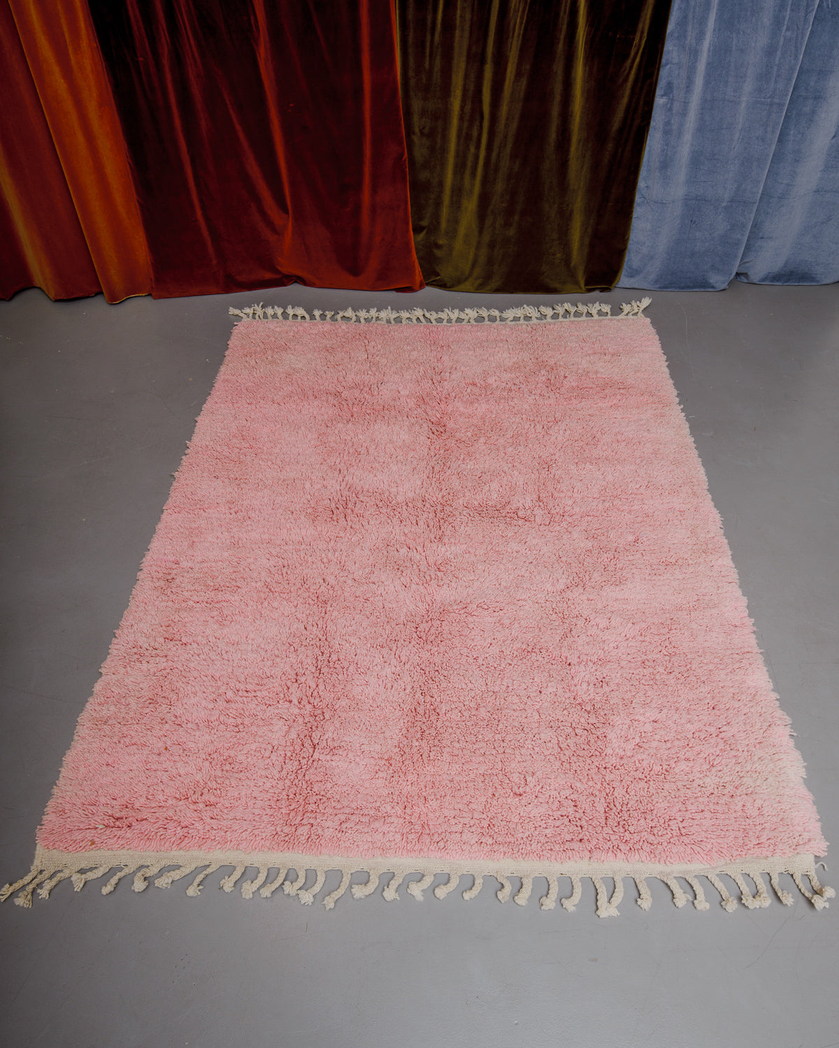 Berberlin Boujaad rug in soft rose wool with natural wool tassels at both ends on grey floor in front of velvet curtains.