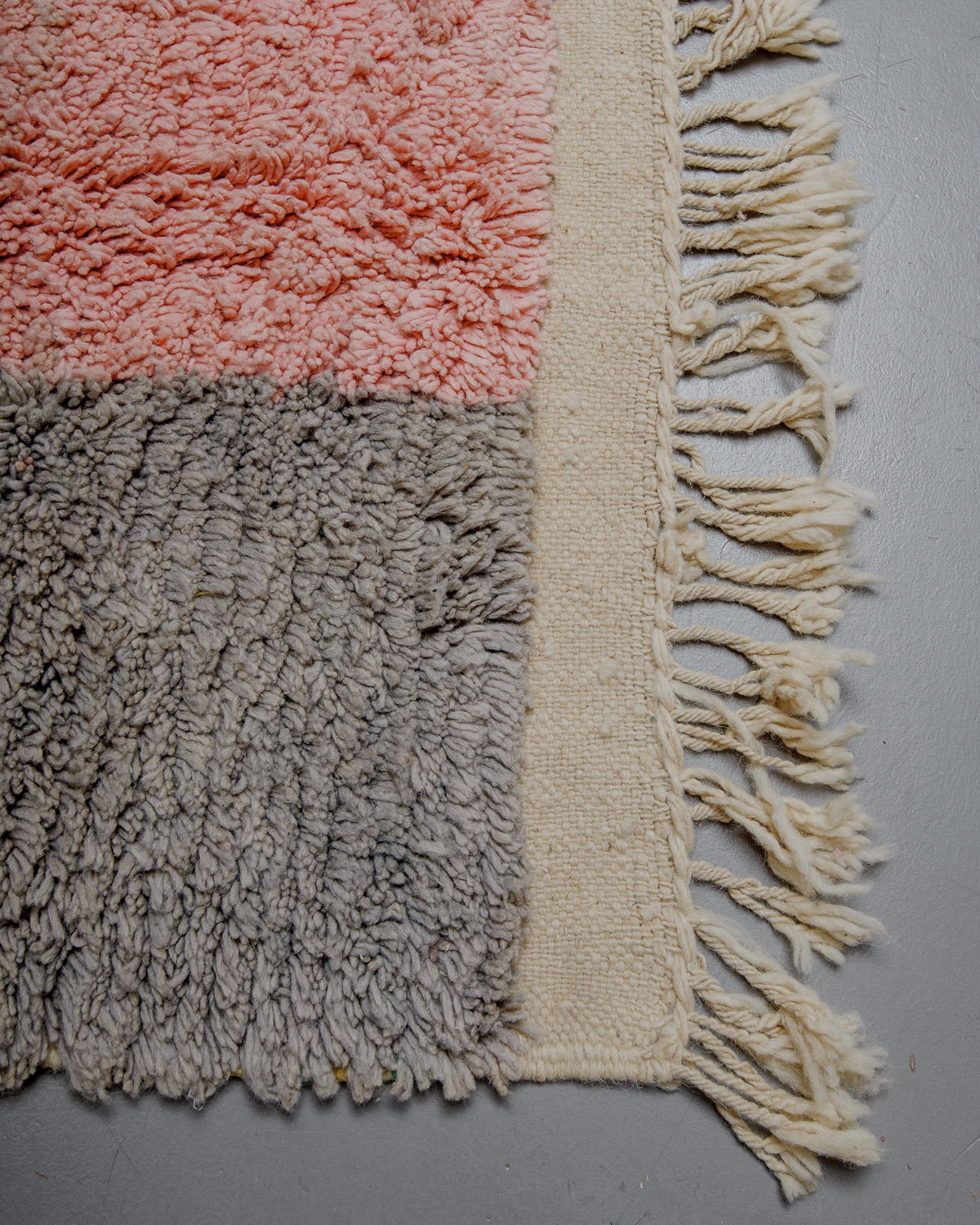 Corner of wool rug with pink and grey pile and flat woven edge with natural wool tassels. 