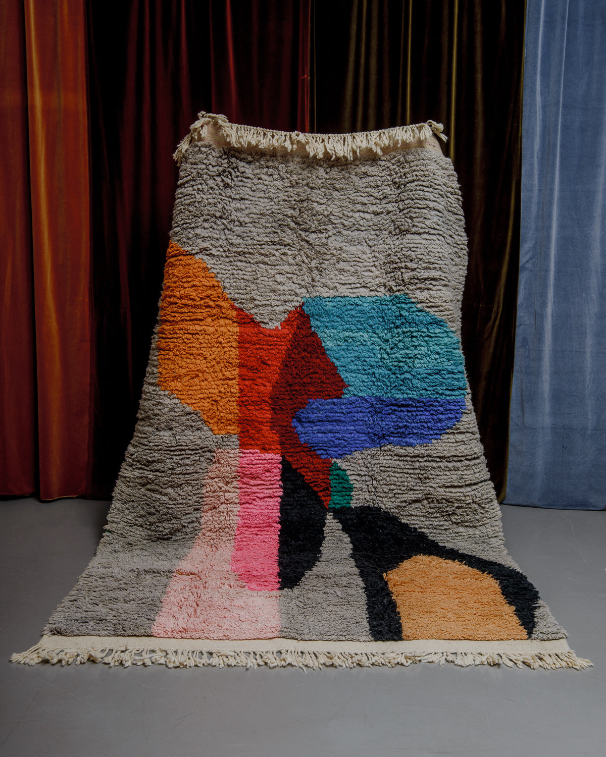 Berberlin Boujaad rug with bold forms in orange, red, blue, pink and grey being held in front of velvet curtains.