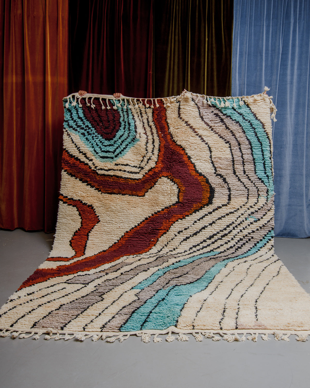 Berberlin Boujaad rug with bold, organic forms and lines in beige, grey, rust and turquoise being held in front of velvet curtains.