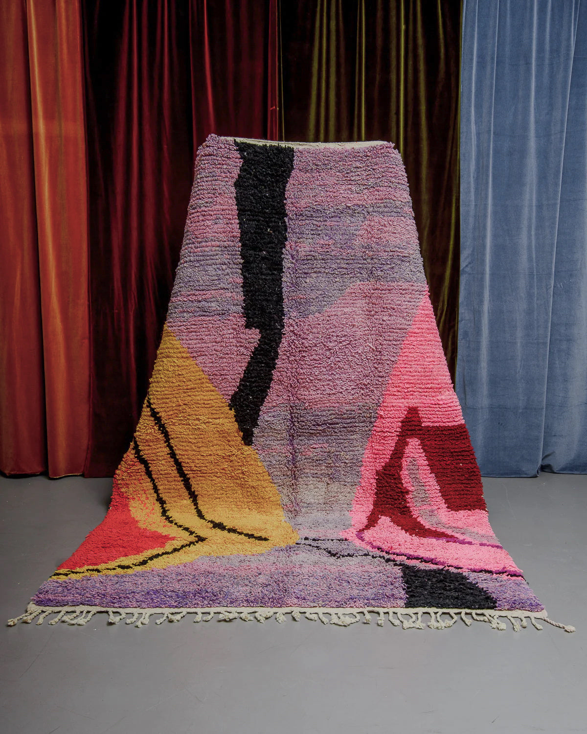 Berberlin Boujaad rug with bold, organic forms and lines in lavender, pink, yellow and red being held in front of velvet curtains.
