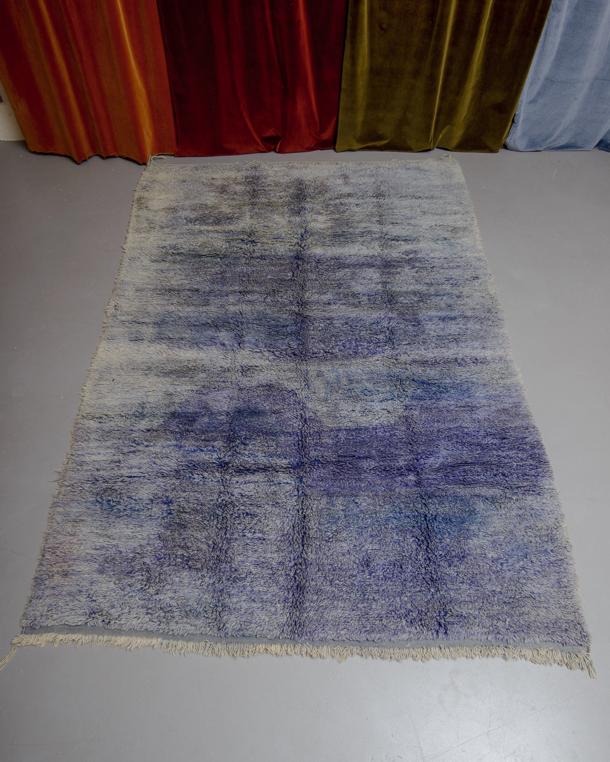 Berberlin Boujaad rug in marled blue and purple wool on grey floor in front of velvet curtains.