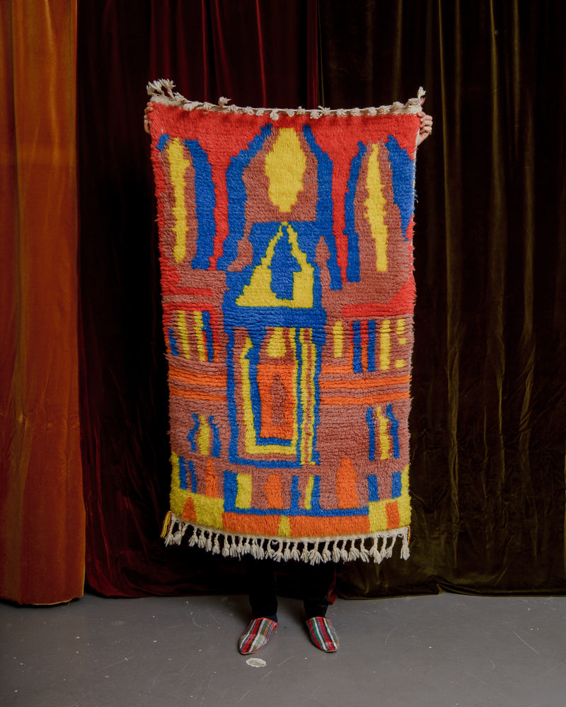 Colourful rug being held up in front of a wall of velvet curtains.