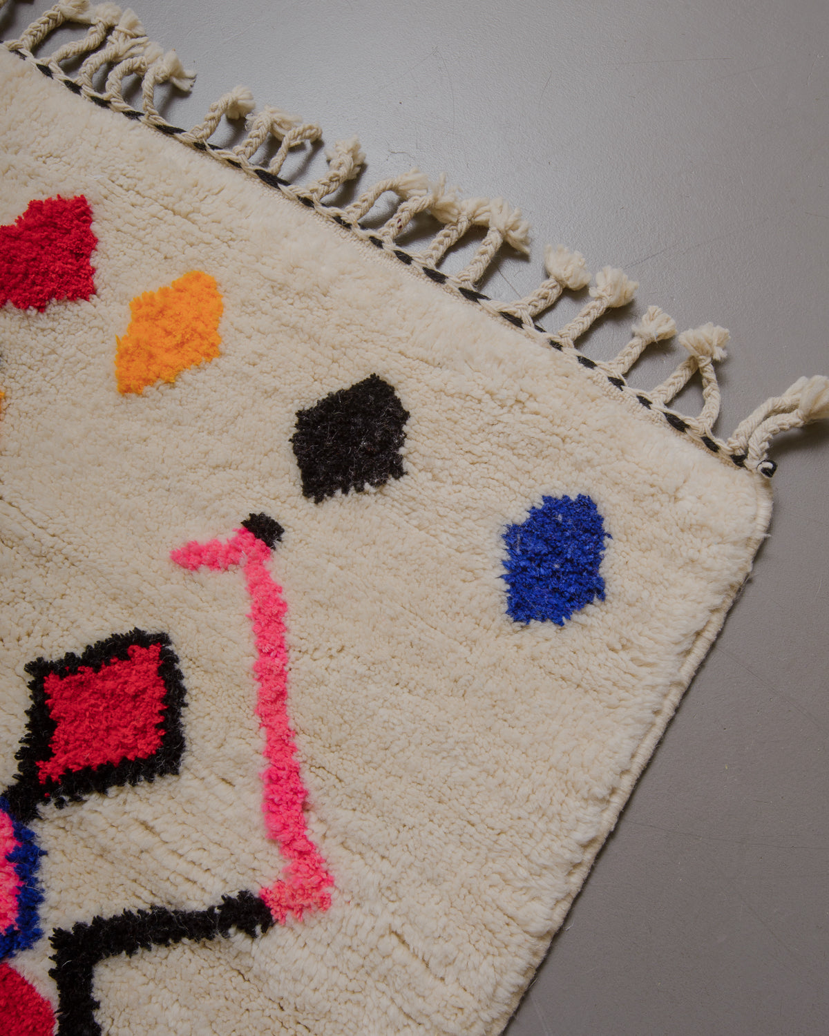 Corner of cream rug with colourful elements and natural wool tassels on a grey floor. 