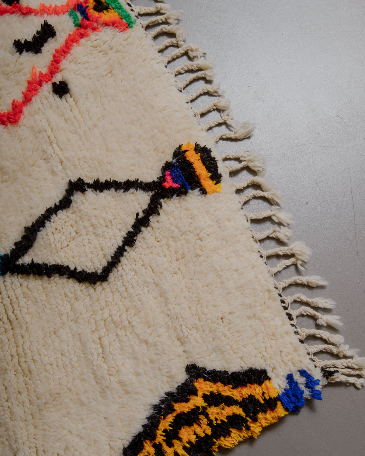 Corner of cream rug with colourful elements and natural wool tassels on a grey floor. 