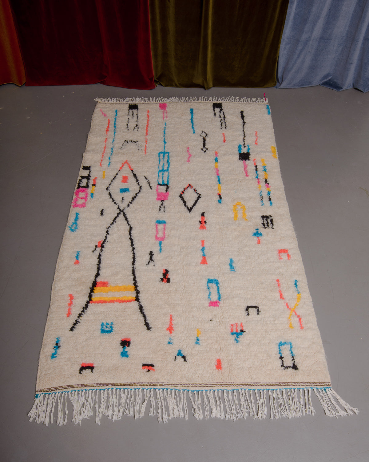 Azilal rug with colourful, abstract design laid flat on grey floor in front of a wall of velvet curtains.