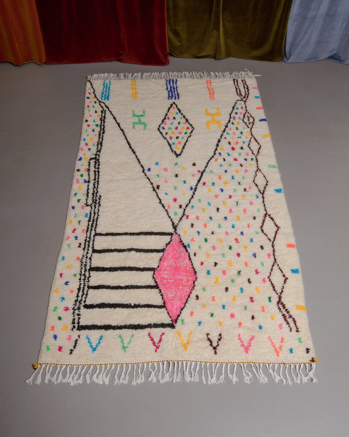 Azilal rug with colourful, abstract design laid flat on grey floor in front of a wall of velvet curtains.