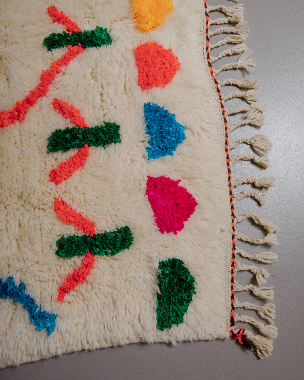 Corner of cream rug with colourful elements and natural wool tassels on a grey floor. 