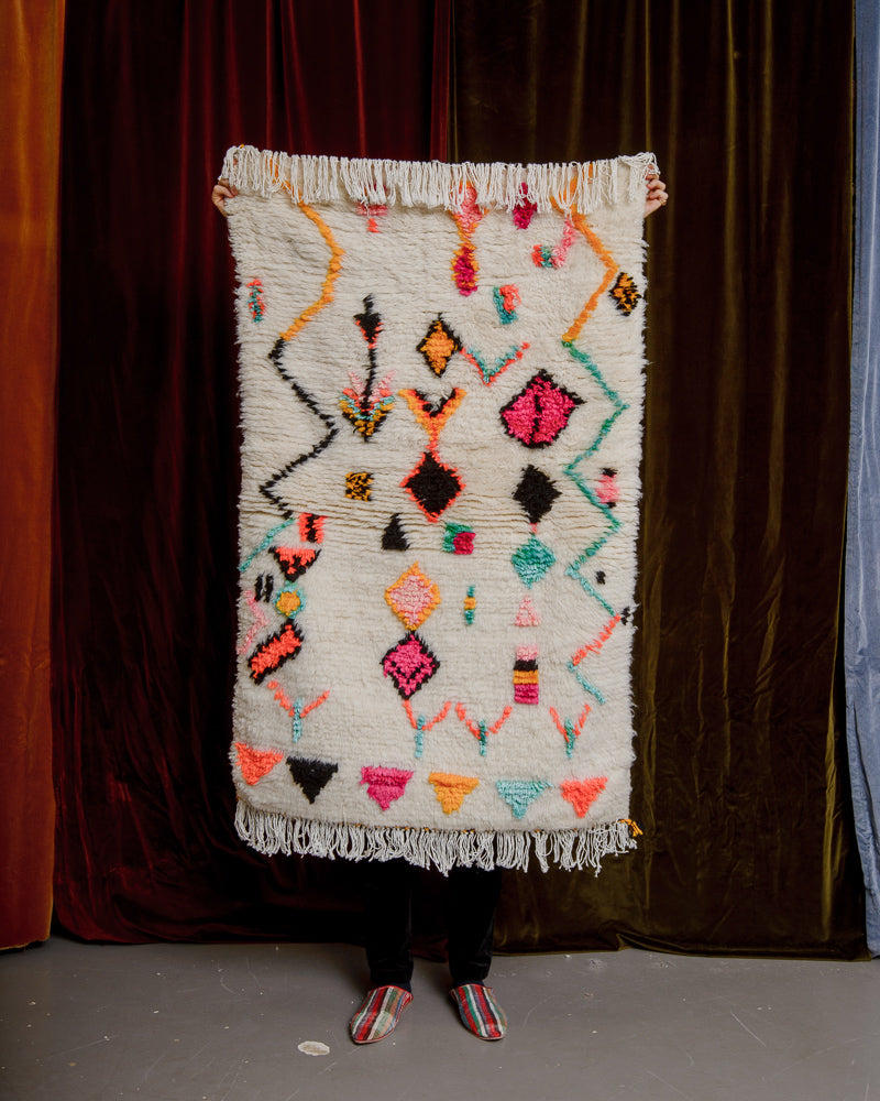 Brightly coloured Berberlin azilal rug in a small size, being held up in front of a velvet curtain.