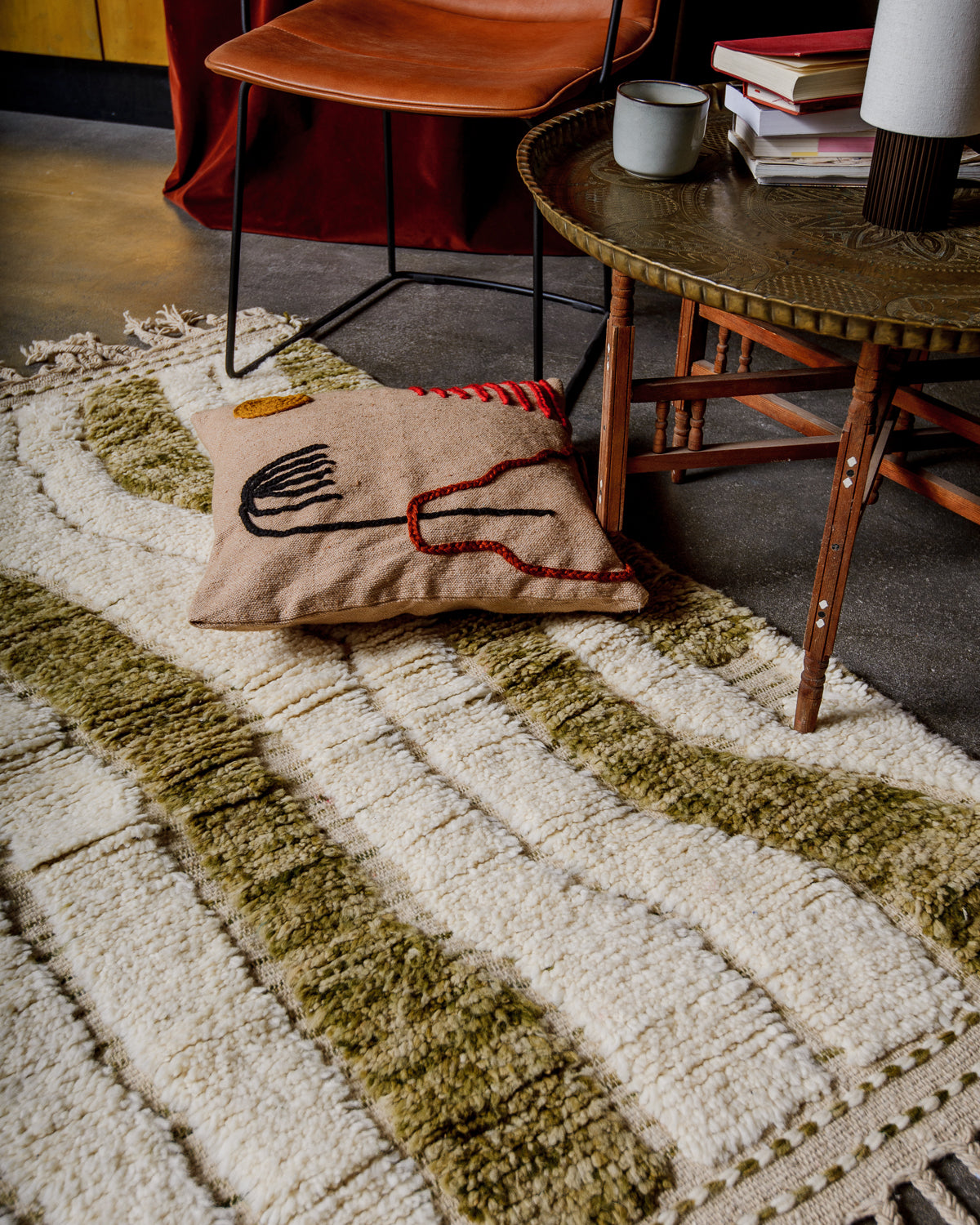 Interior shot of small Beniouran rug in cream and khaki with cushion, chair and small brass table. 